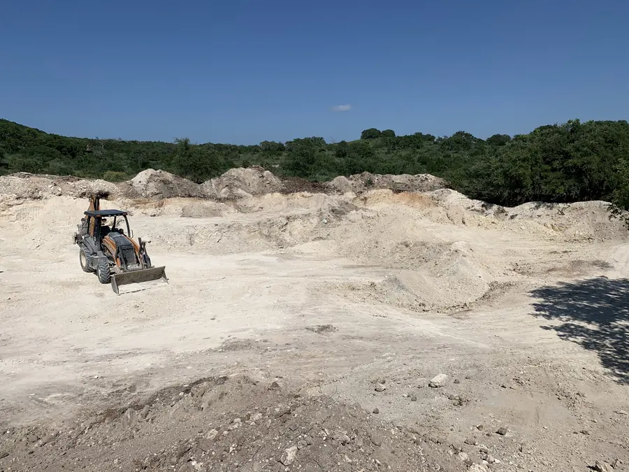 Backhoe loader parked in a wide pale rock and caliche dig, with spoil piles and green brush behind